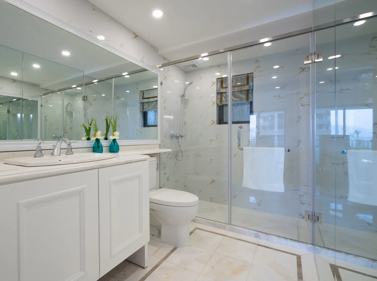 Frameless glass shower enclosure with chrome hinges in a bright marble-tiled bathroom.