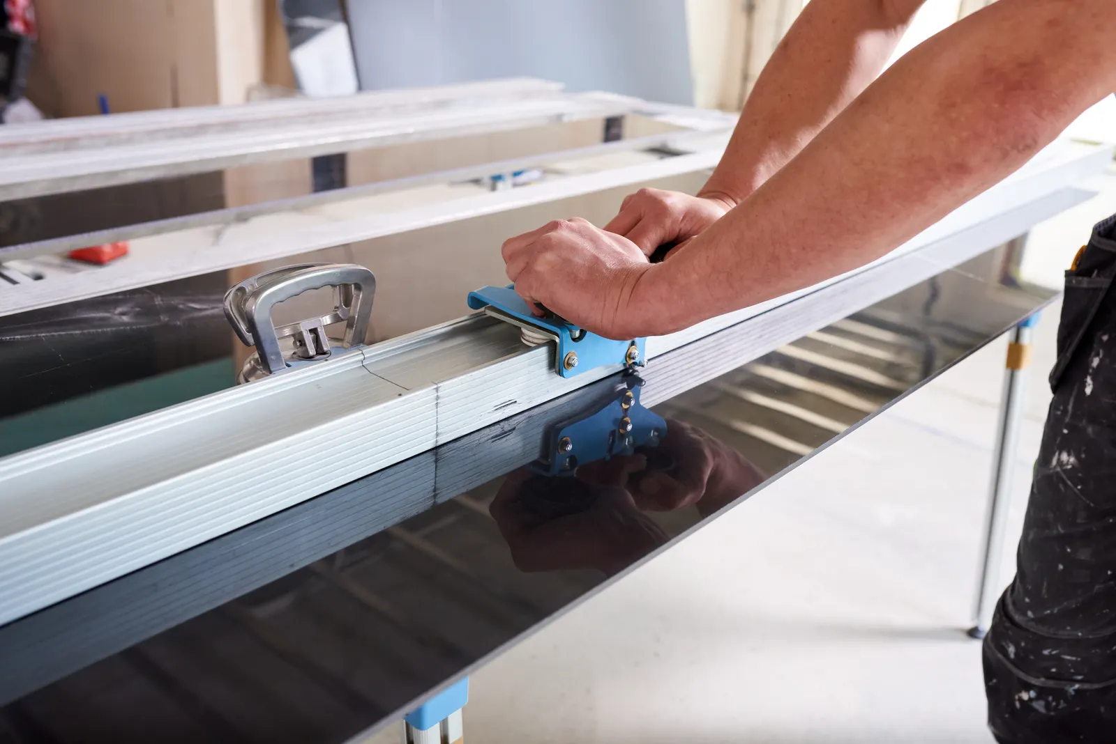 A worker cutting a large dark glass panel with a straightedge tool on a workbench.