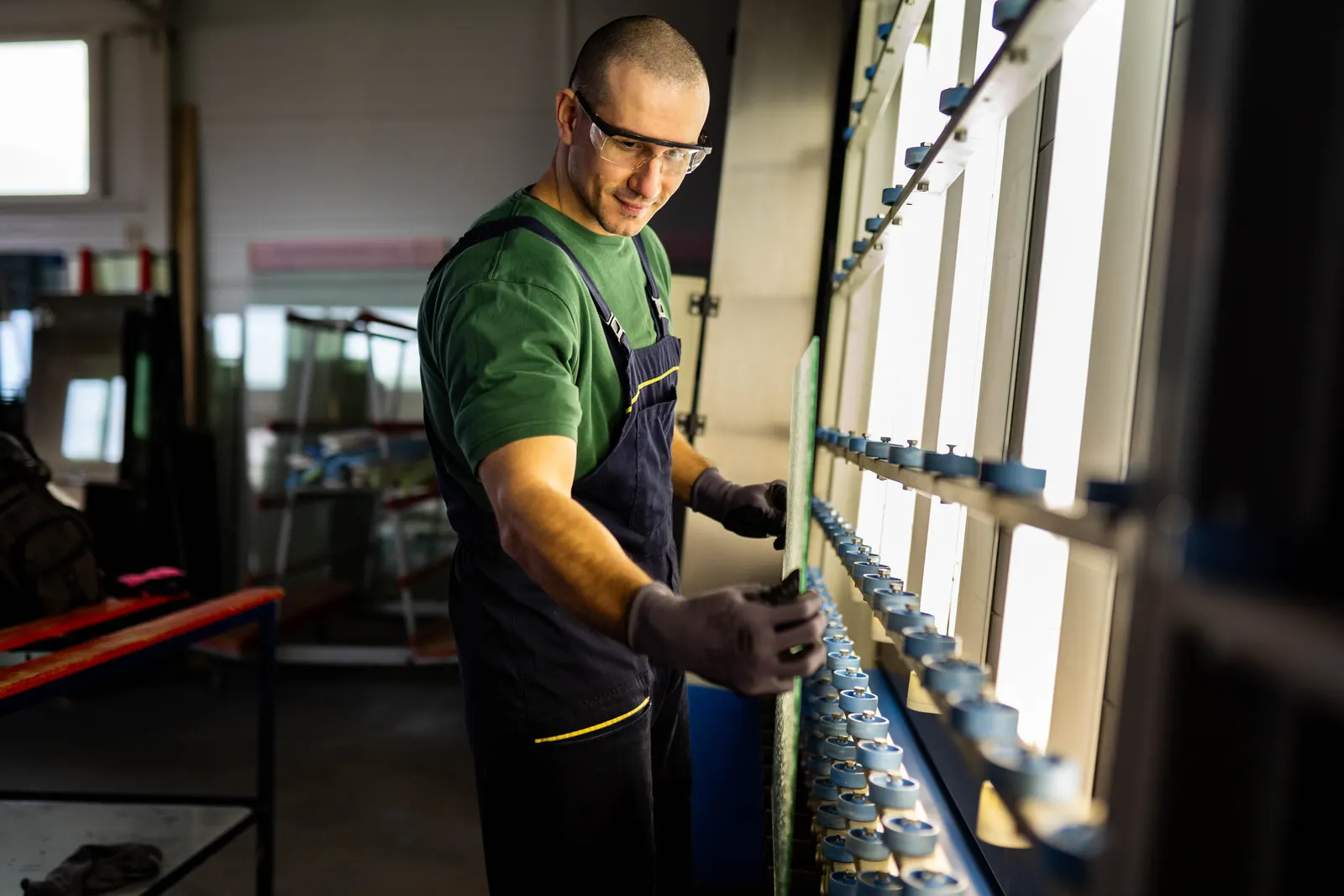 A worker in safety glasses feeding a glass sheet into a roller machine in a workshop.