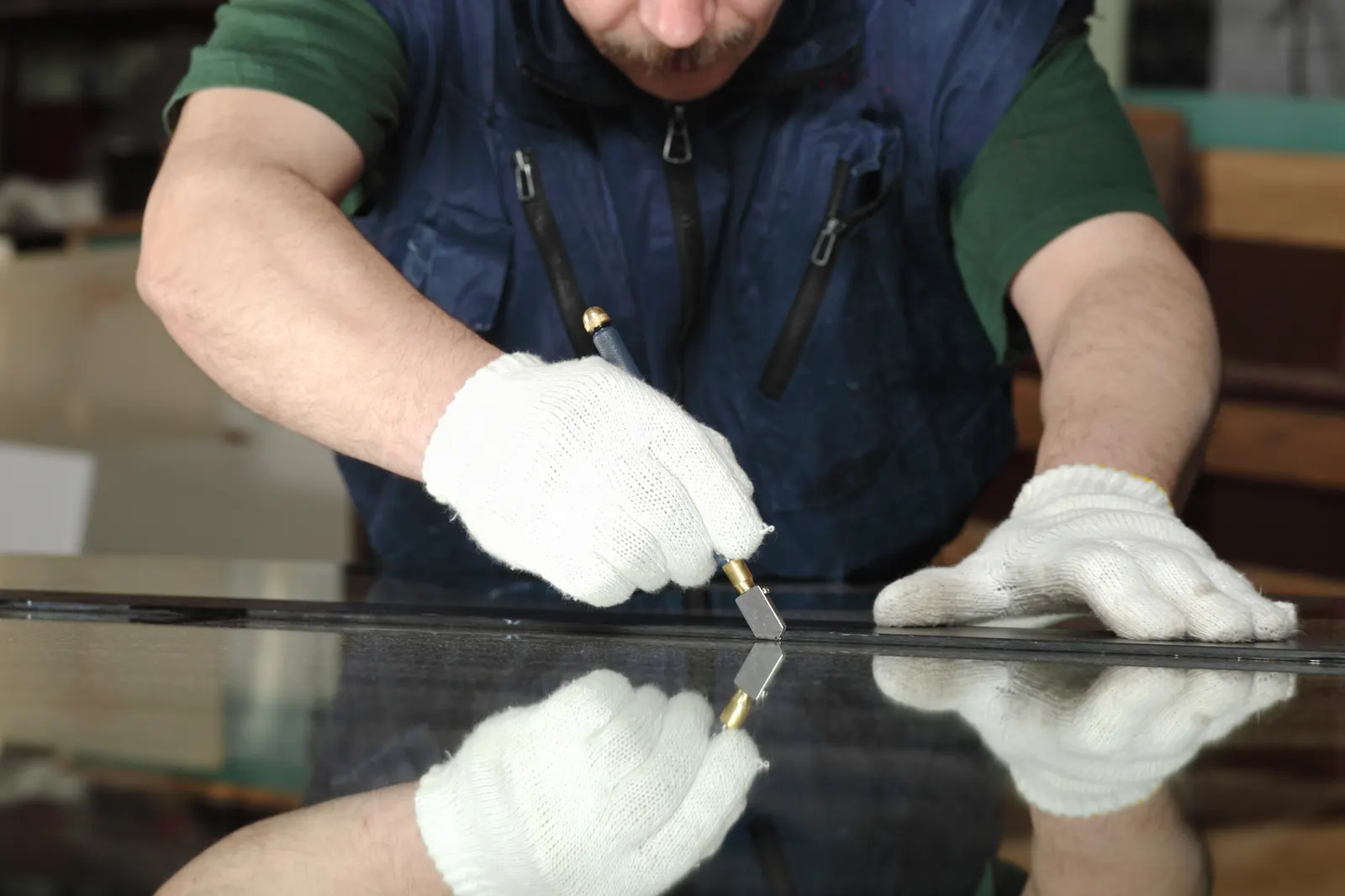 A worker in white gloves scoring a dark glass pane with a handheld glass cutter.