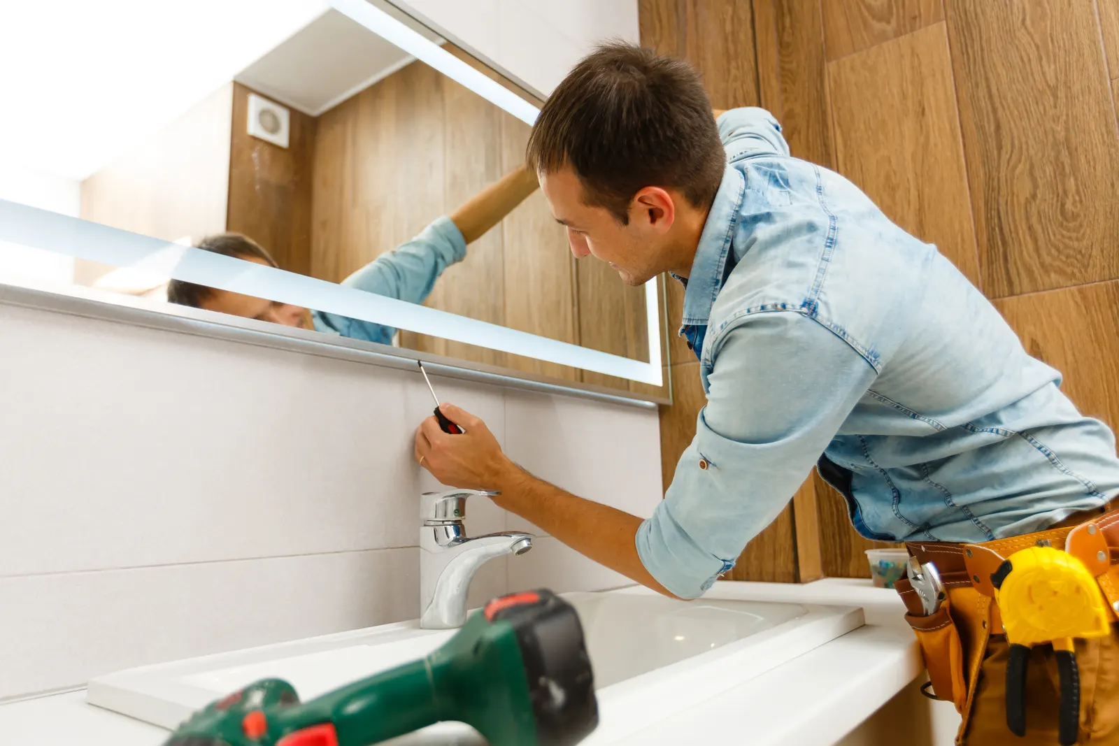 A worker installing a large backlit frameless bathroom mirror above a sink.