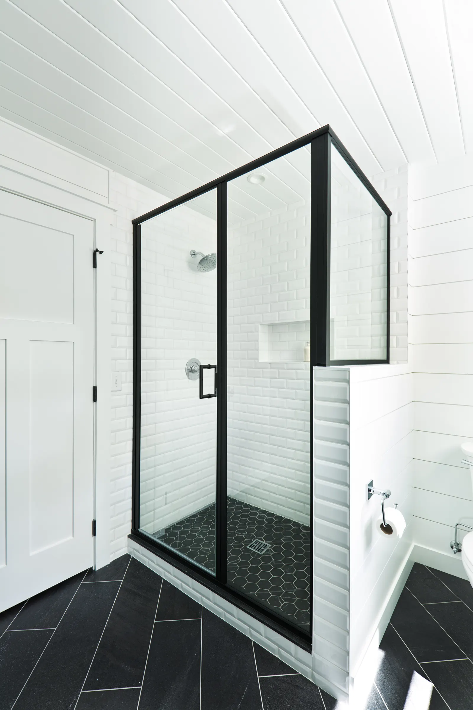 Black-framed hinged glass shower door with white subway tile and a black hexagon floor.