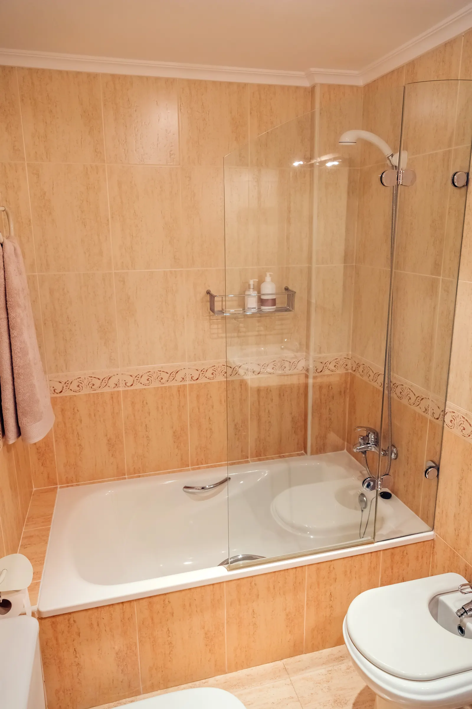 Bathtub with a folding frameless glass bath screen and beige travertine tile.