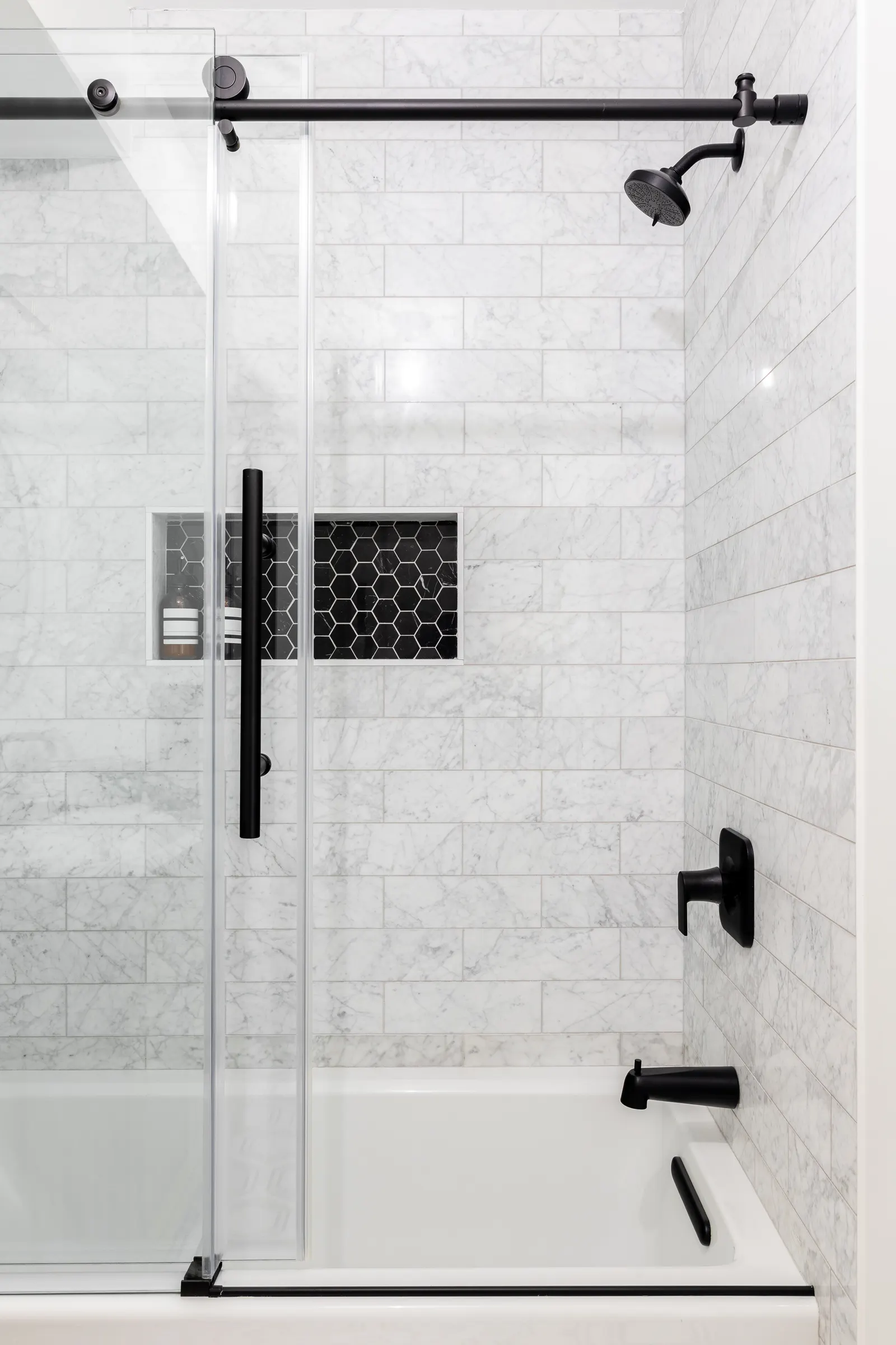 Bathtub with a frameless sliding glass tub door on a black rail and marble tile.