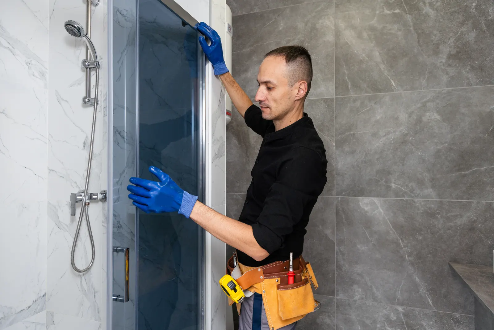 A worker in gloves and a tool belt adjusts a sliding glass shower door on marble tile.