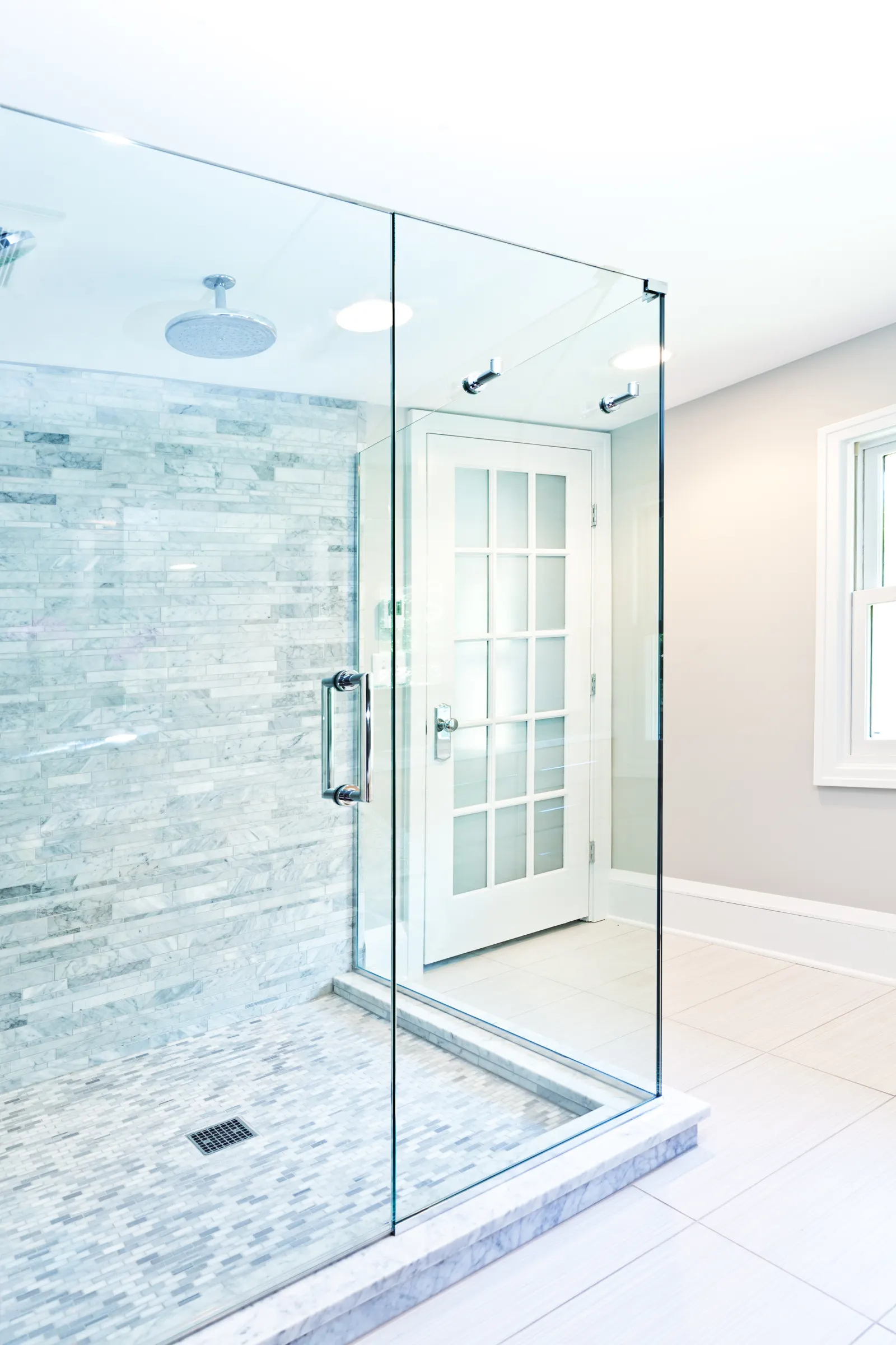 Frameless glass shower enclosure with a chrome handle and marble tile beside a white French door.