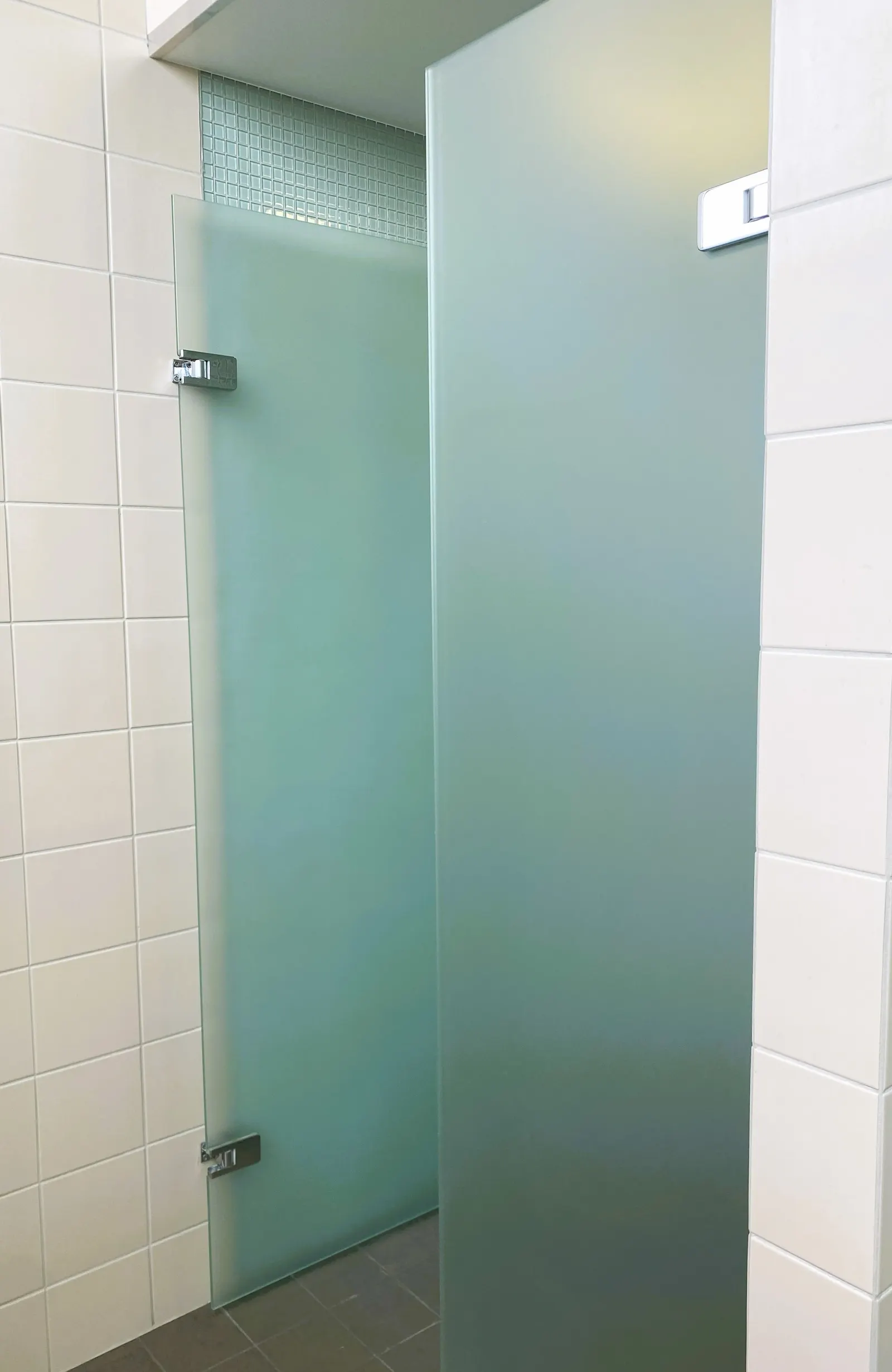 Open frosted glass shower door on chrome hinges beside white-tiled walls.