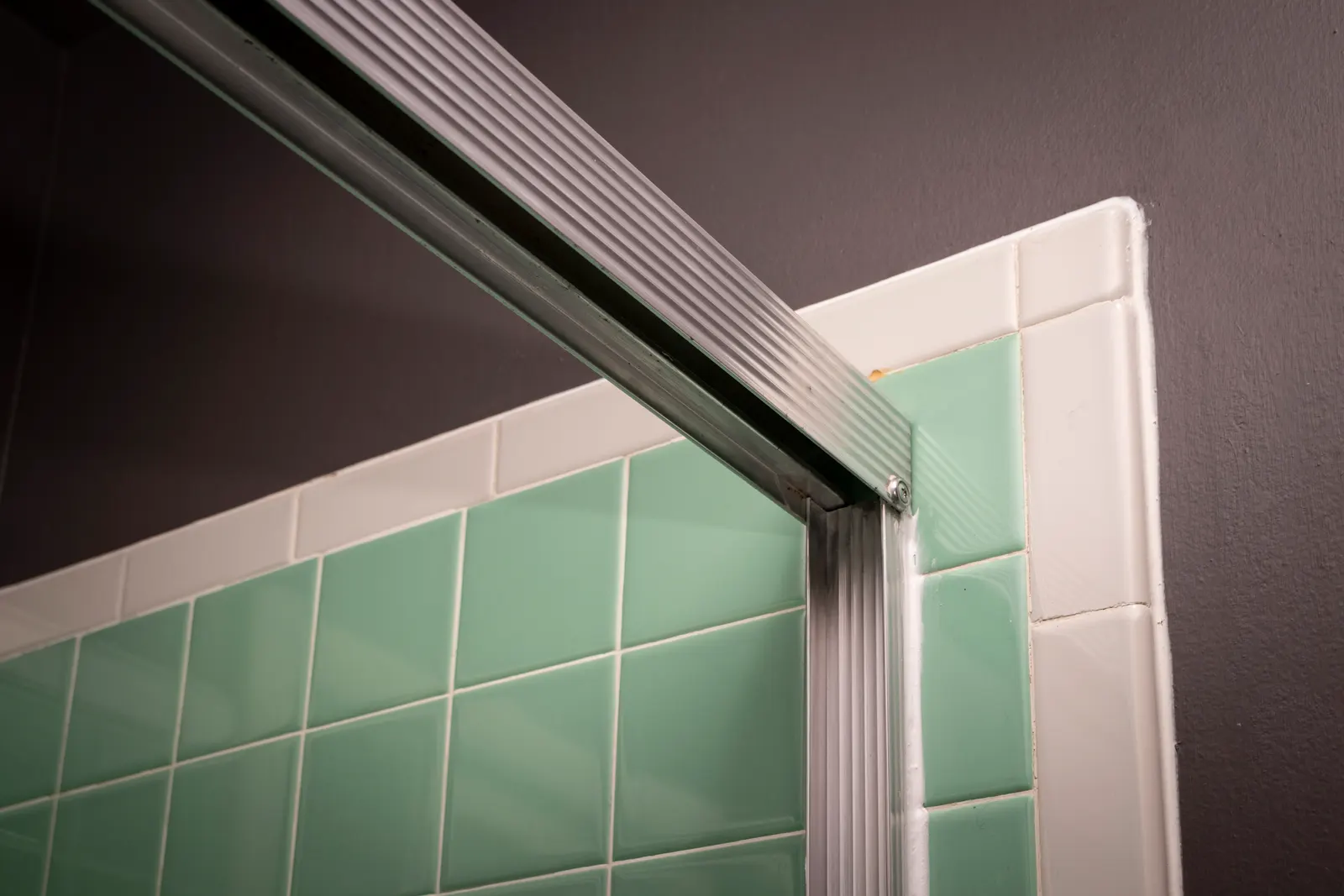 Close-up of an aluminum framed sliding shower door track above green wall tile.