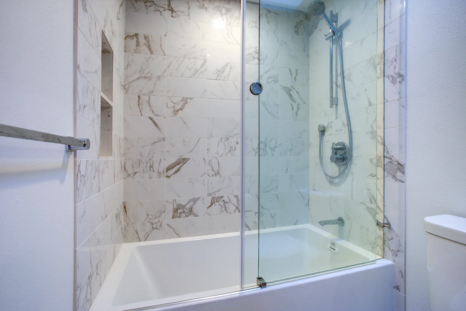 Bathtub with a frameless pivoting glass bath screen and white marble tile.