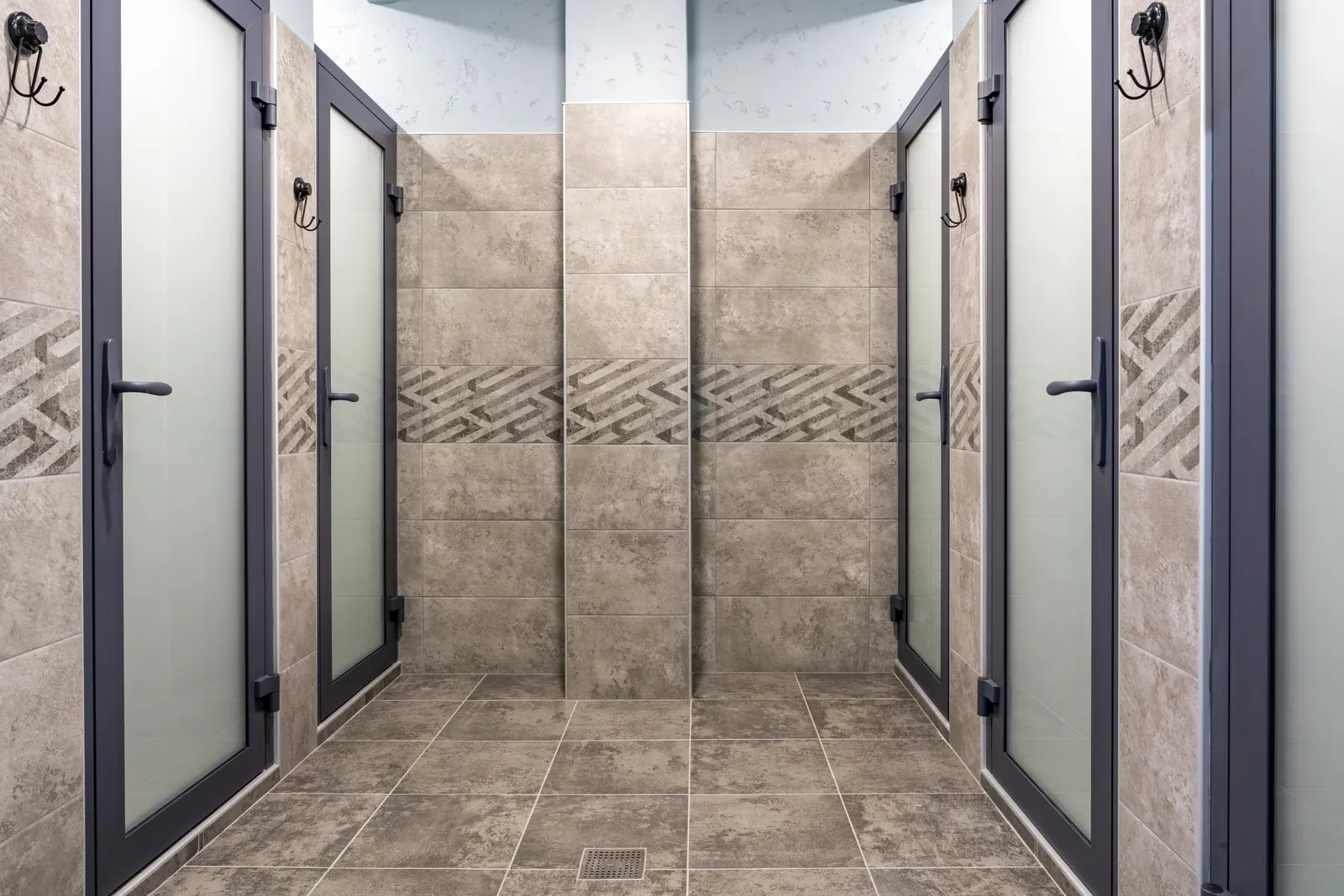 Row of tiled shower stalls with frosted glass doors and dark frames in a locker room.