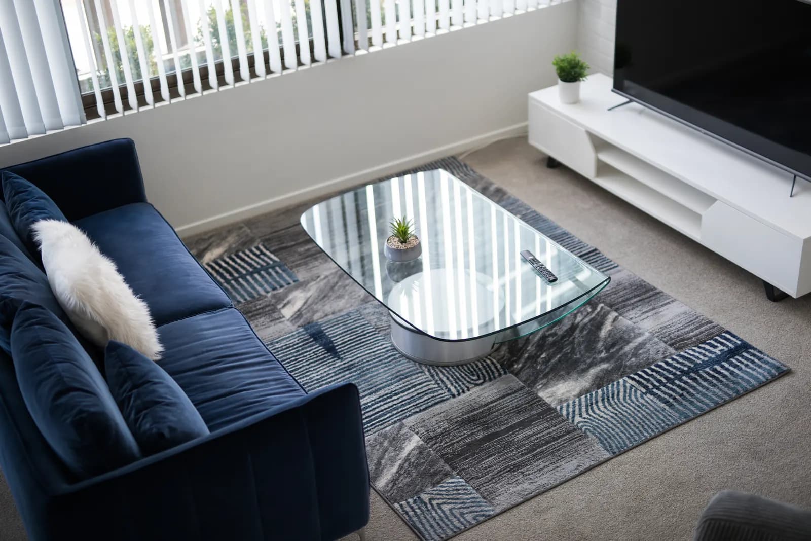 Rounded glass-top coffee table on a patterned rug beside a blue velvet sofa in a living room.