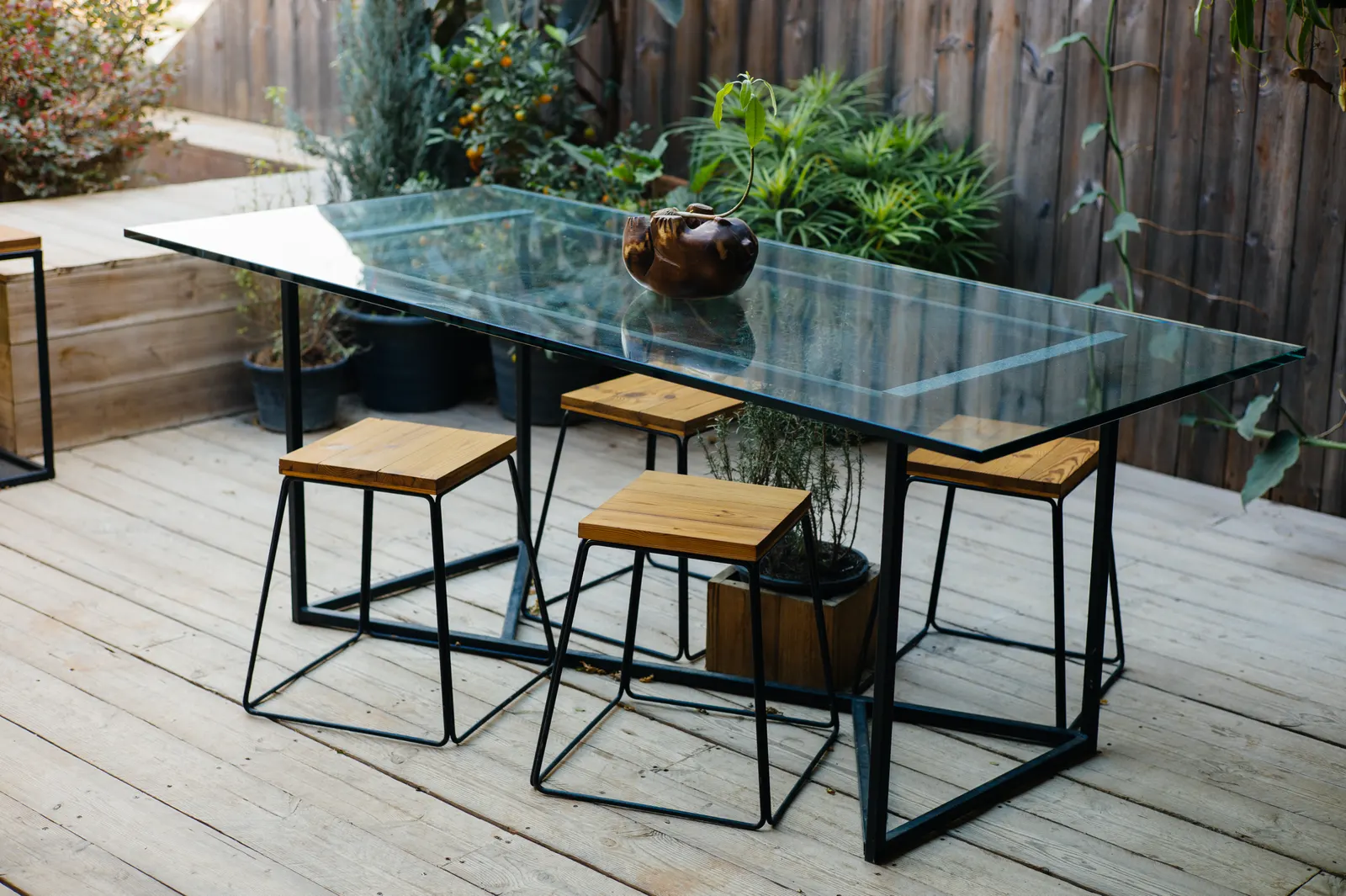 Outdoor dining table with a clear glass top on a black metal frame and wooden stools.