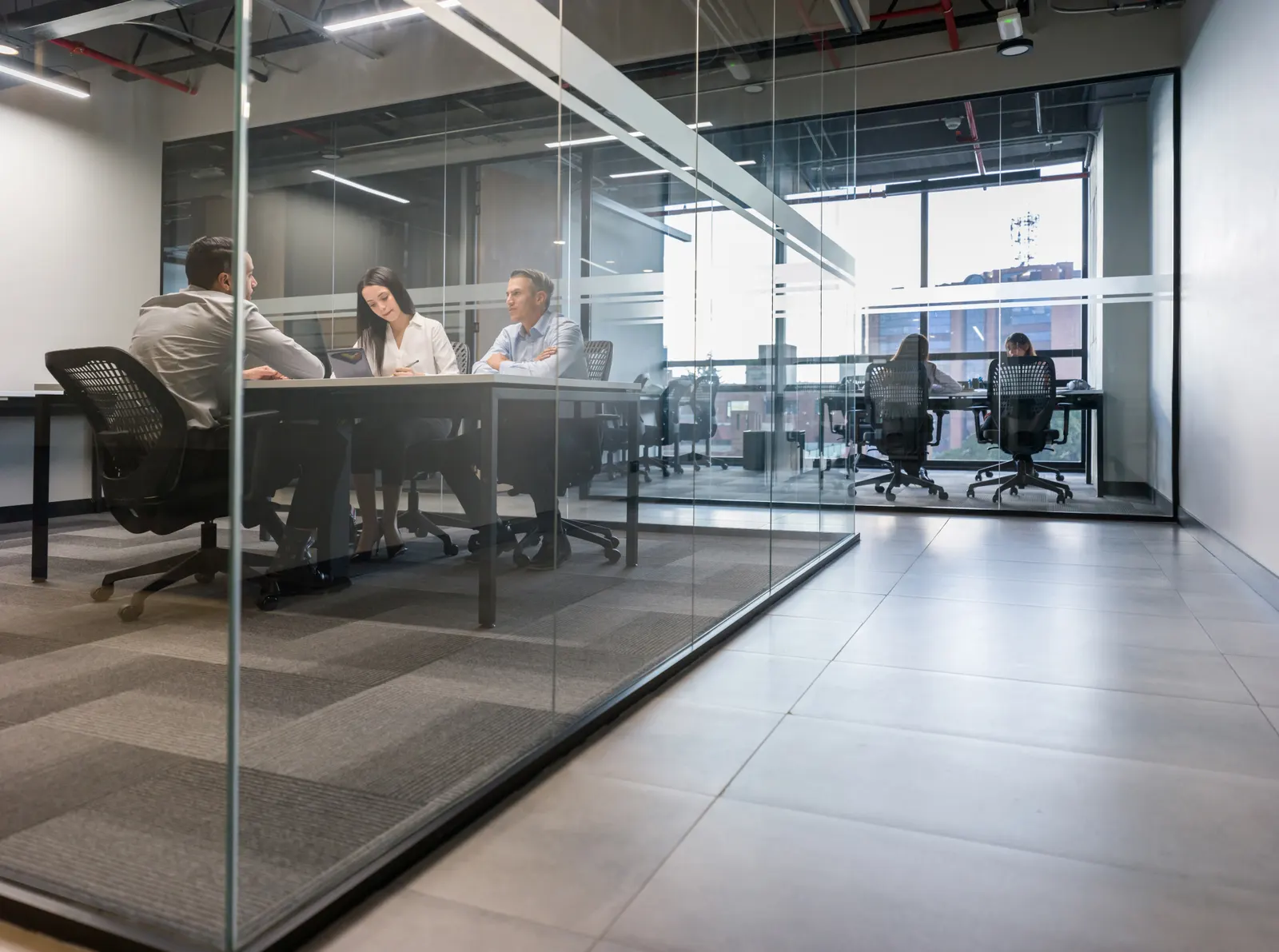 Colleagues seated in a glass-walled meeting room beside an open office.