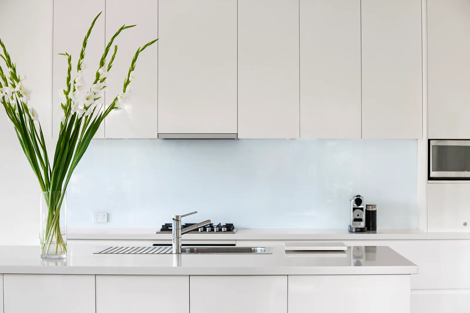 White glass backsplash behind a cooktop and sink in a white kitchen.